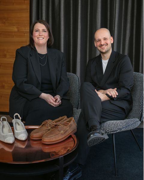 Zwei Personen in schwarzen Anzügen sitzen lächelnd auf Stühlen, vor ihnen stehen zwei Paar Schuhe auf einem runden Tisch. Im Hintergrund ist ein dunkler Vorhang zu sehen.