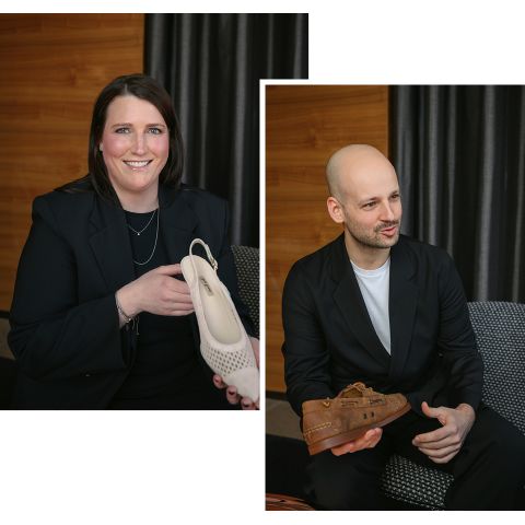 Eine Frau hält einen beigen Schuh und lächelt, ein Mann hält einen braunen Schuh und schaut zur Seite. Beide sind in dunkle Blazer gekleidet und sitzen in einem Haus.