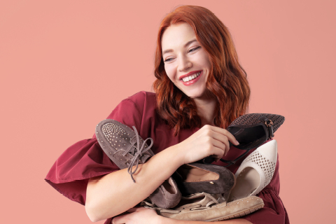 Lächelnde Frau mit roten Haaren in einem kastanienbraunen Oberteil, die mehrere Paar Schuhe vor einem pfirsichfarbenen Hintergrund hält.