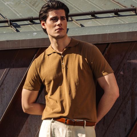 Ein Mann in einem braunen Polohemd und einer beigen Hose steht im Freien im Sonnenlicht, eine Hand in der Tasche, vor einer Metallkonstruktion.