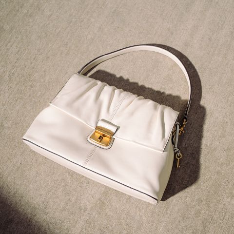 Cremefarbene Handtasche mit goldener Schnalle und einfachem Henkel, die auf einer hellen Betonfläche in natürlichem Sonnenlicht ruht.