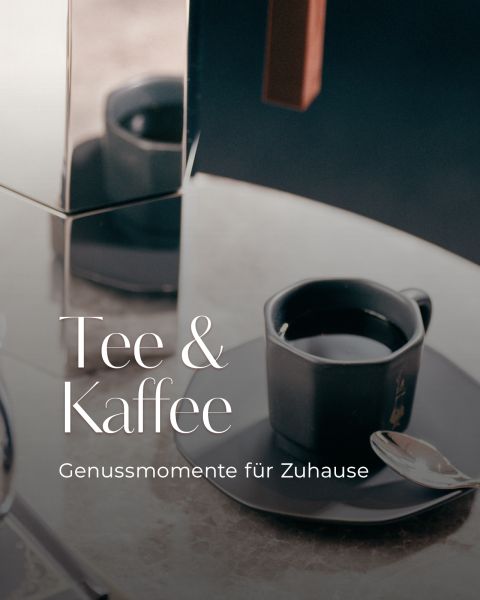 Eine schwarze Tasse Kaffee auf einer Untertasse mit einem Löffel, neben einer spiegelnden Oberfläche. Text: "Tee & Kaffee - Genussmomente für Zuhause.