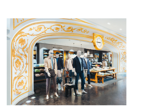 Eingang eines Bekleidungsgeschäfts mit verschnörkeltem Goldbogen, Schaufensterpuppen in Freizeitkleidung und ordentlich gefaltete Kleidung in Regalen und auf Tischen.