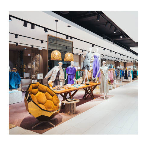 Ein Bekleidungsgeschäft mit Schaufensterpuppen in modischen Outfits, Holztischen mit Accessoires und einem einzigartigen goldenen, geometrischen Stuhl im Vordergrund.