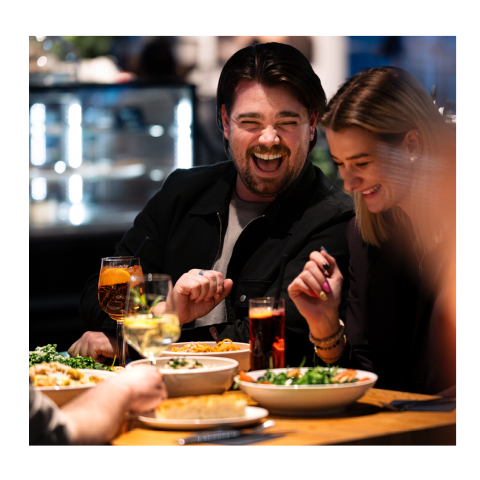 Zwei Menschen lachen und genießen eine Mahlzeit in einem Restaurant, umgeben von Getränken und Tellern mit Speisen auf einem Holztisch.