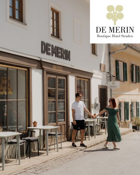 Ein Paar hält sich an den Händen, während es vor dem De Merin Boutique Hotel Straden spazieren geht, mit Sitzgelegenheiten im Freien und sonnenbeschienenen Gebäuden im Hintergrund.