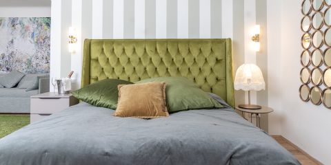 Ein stilvolles Schlafzimmer mit einem grünen getufteten Kopfteil, grauer Bettwäsche, grünen und goldenen Kissen, Nachttischen, Lampen und dekorativen Wandspiegeln.