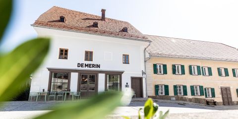 Straßenansicht eines weißen Gebäudes mit "DEMERIN"-Schild, Außenbestuhlung, neben einem gelben Haus mit grünen Fensterläden, unter klarem Himmel.