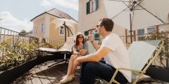 Ein Mann und eine Frau entspannen und unterhalten sich auf einer sonnigen Dachterrasse mit Liegestühlen, Pflanzen und großen weißen Sonnenschirmen.