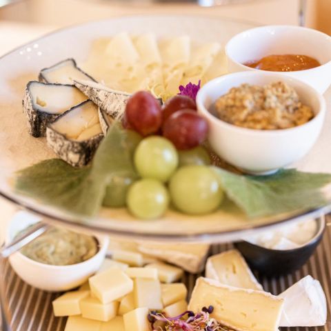 Eine Platte mit verschiedenen Käsesorten, grünen und roten Weintrauben, Obstkonserven und einer kleinen Schale mit Nüssen, angerichtet auf einem weißen Teller.