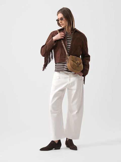 Frau in brauner Wildlederjacke mit Fransen, gestreiftem Hemd, weißer Hose mit weitem Bein, braunen Schuhen, Sonnenbrille und hellbrauner Umhängetasche, steht selbstbewusst da.