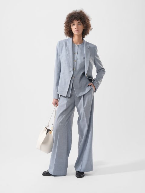 Eine Frau mit lockigem Haar trägt einen locker sitzenden hellgrauen Anzug, schwarze Schuhe und hält eine weiße Handtasche vor einem schlichten weißen Hintergrund.