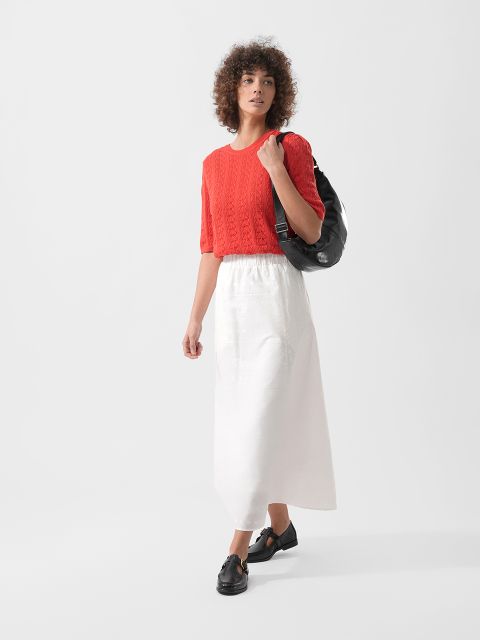 Frau mit lockigem Haar, die einen roten kurzärmeligen Pullover, einen weißen knöchellangen Rock und schwarze Schuhe trägt und eine schwarze Tasche über der Schulter hat.