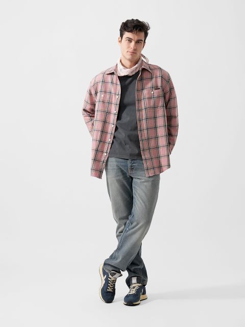 Stehender Mann mit Händen in den Taschen, der ein rosa kariertes Hemd über einem grauen T-Shirt, helle Jeans, marineblaue Turnschuhe und einen hellen Schal trägt, vor einem weißen Hintergrund.