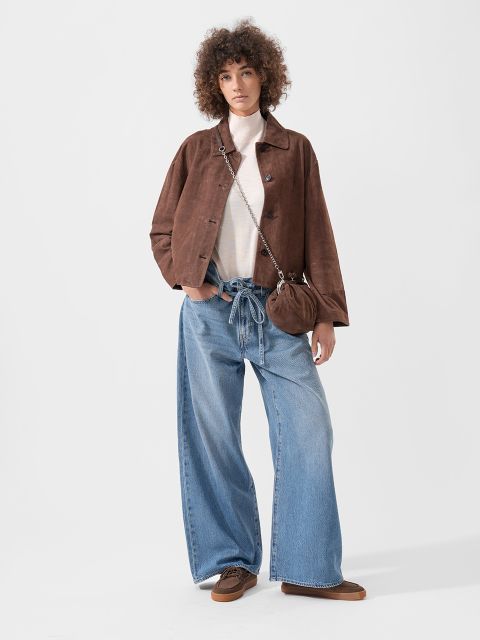 Person, die eine braune Jacke, ein weißes Oberteil, eine weit geschnittene blaue Jeans, braune Schuhe und eine kleine braune Handtasche trägt und vor einem schlichten Hintergrund steht.