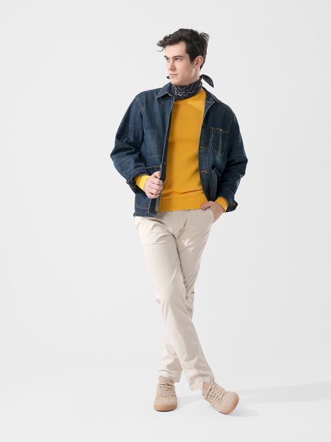 Ein Mann in einem gelben Pullover, Jeansjacke, beiger Hose und hellbraunen Schuhen posiert mit einer Hand in der Tasche vor einem schlichten weißen Hintergrund.