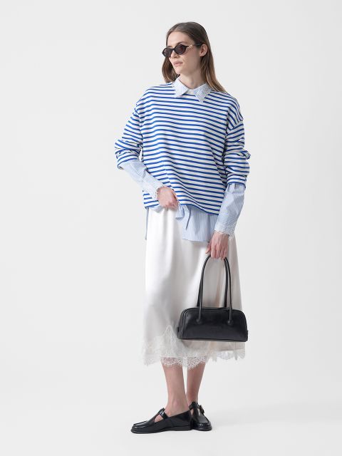 Frau mit Sonnenbrille, die einen blau-weiß gestreiften Pullover über einem Hemd mit Kragen, einen weißen Rock mit Spitzenbesatz, schwarze Halbschuhe und eine schwarze Handtasche trägt.