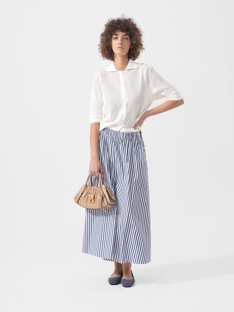 Frau in einem weißen kurzärmeligen Strickoberteil, einem blau-weiß gestreiften Maxirock, marineblauen Flats und einer beigen Handtasche vor einem schlichten Hintergrund.