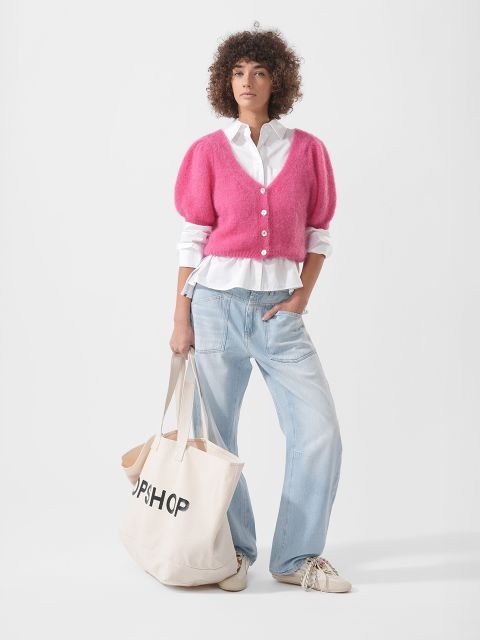 Eine Person in rosa Strickjacke, weißem Hemd, hellblauen Jeans und Turnschuhen hält eine große beigefarbene Tragetasche mit dem Aufdruck "TOPSHOP" in der Hand.