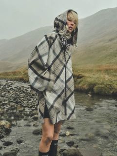 Eine Person, die einen karierten Regenmantel mit Kapuze trägt, steht an einem felsigen Bach in einer nebligen, grasbewachsenen Landschaft, mit Hügeln im Hintergrund.