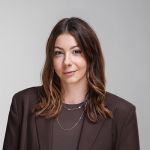 Eine Frau mit gewelltem braunem Haar, die einen braunen Blazer und mehrlagige Halsketten trägt, posiert vor einem schlichten hellgrauen Hintergrund.