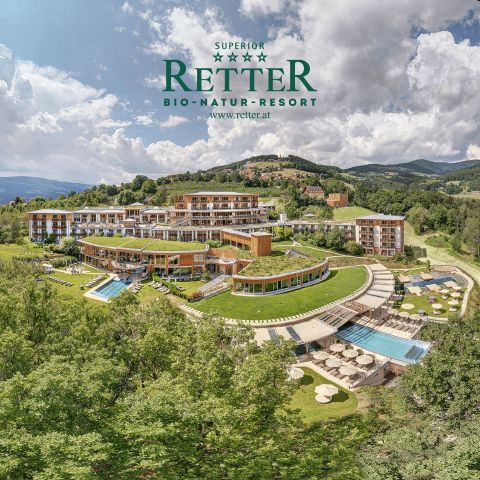 Luftaufnahme des RETTER Bio-Natur-Resorts inmitten von Grünanlagen, mit Pools, Terrassen und Hügeln im Hintergrund unter einem teilweise bewölkten Himmel.