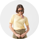 Frau, die einen gelben kurzärmeligen Pullover, eine olivgrüne Hose, eine Sonnenbrille und ein gemustertes Halstuch trägt und vor einem hellgrauen Hintergrund steht.