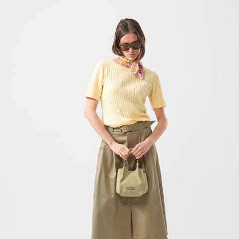 Frau mit Sonnenbrille und gelbem, kurzärmeligem Oberteil mit Schal, khakifarbenem Rock und einer kleinen grünen Tasche in der Hand vor einem schlichten weißen Hintergrund.
