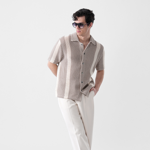 Mann mit Sonnenbrille, beigefarbenem, gemustertem, kurzärmeligem Hemd und heller Hose, der mit einer Hand in der Tasche vor einem schlichten Hintergrund steht.