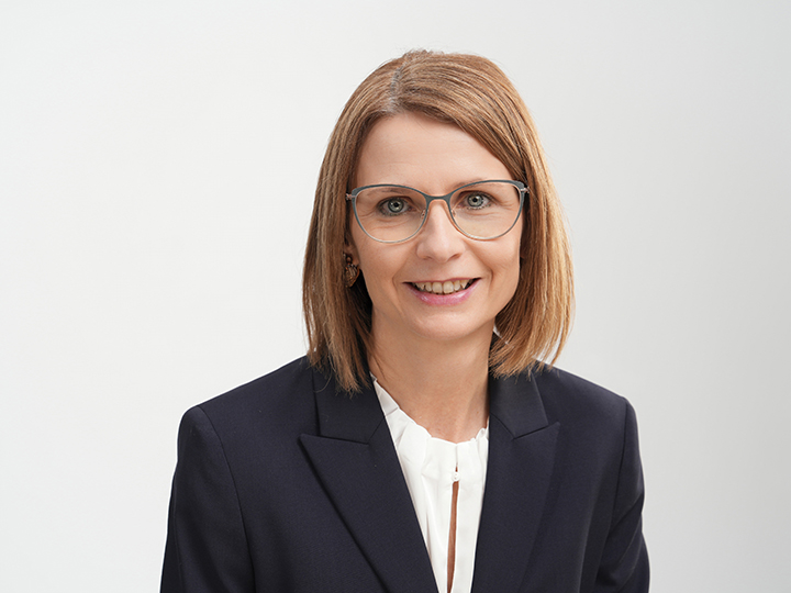 Frau mit glattem, schulterlangem hellbraunem Haar, die eine Brille, einen marineblauen Blazer und eine weiße Bluse trägt und vor einem schlichten hellen Hintergrund lächelt.