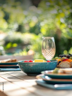 Eine türkisfarbene Schale mit Obst steht auf einem gedeckten Tisch mit Tellern, Brot und einem Glas in einem sonnenbeschienenen Garten.
