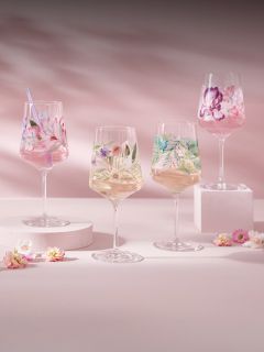 Vier Weingläser mit bunten Blumenmustern, gefüllt mit Getränken, stehen auf pastellfarbenen Podesten mit verstreuten kleinen Blumen auf einem rosa Hintergrund.