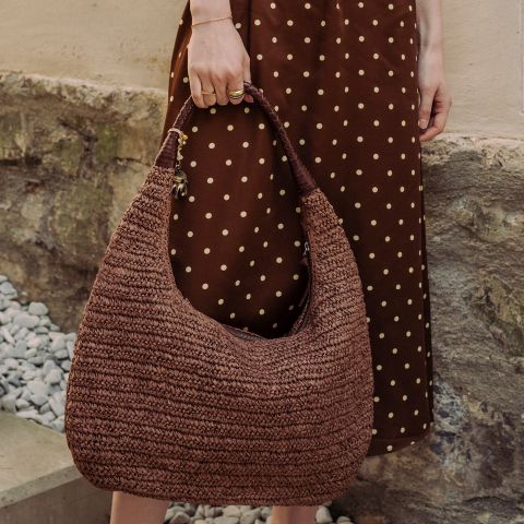 Eine Person in einem braunen gepunkteten Kleid hält eine große, braune gewebte Handtasche in der Hand, während sie neben einer Steinmauer und Kies steht.