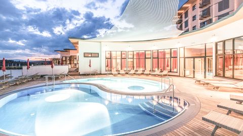 Ein moderner Außenpoolbereich mit runden Becken, Liegestühlen und großen Fenstern, die bei Sonnenuntergang mit einem dramatischen Himmel im Hintergrund beleuchtet werden.