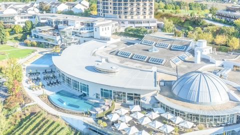 Luftaufnahme eines modernen Resorts mit Pool, Liegestühlen, kuppelförmigem Gebäude und angelegten Gärten an einem sonnigen Tag.
