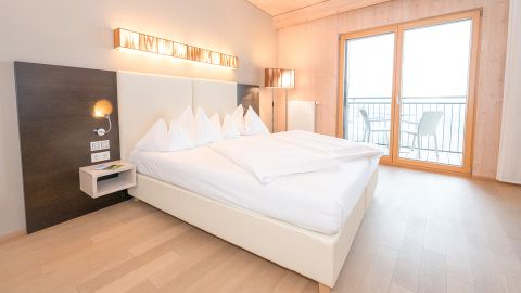 Modernes Hotelzimmer mit sauber gemachtem Doppelbett, Holzfußboden, Wandbeleuchtung und einem durch Glastüren einsehbaren Balkon mit Stühlen.