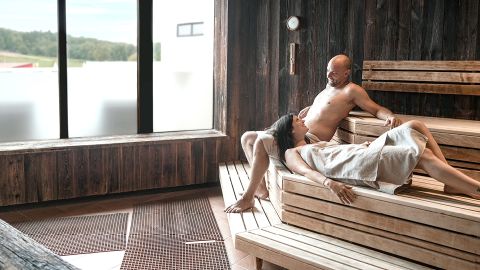 Zwei Personen entspannen sich auf Holzbänken in einer Sauna; eine liegt mit einem Handtuch auf dem Rücken, die andere sitzt aufrecht. Das Sonnenlicht fällt durch große Fenster.