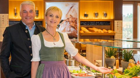 Ein lächelnder Mann und eine lächelnde Frau in traditioneller Kleidung stehen an einer Salatbar in einem gut beleuchteten Restaurant mit Brot und Pflanzen im Hintergrund.