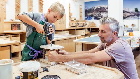 Ein Junge arbeitet mit einer Bohrmaschine an einem Holzprojekt und wird dabei von einem erwachsenen Mann in einem Holzbearbeitungsraum angeleitet.