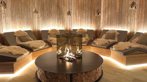 Eine gemütliche Lounge mit Liegesesseln, Felldecken, einer runden Feuerstelle mit Glasabdeckung und warmen, holzgetäfelten Wänden, die von hinten sanft beleuchtet werden.