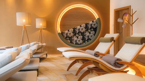 Eine moderne Spa-Lounge mit geschwungenen Liegen, weichen Decken, Stehlampen und einer runden Blumenwand mit warmer Umgebungsbeleuchtung.