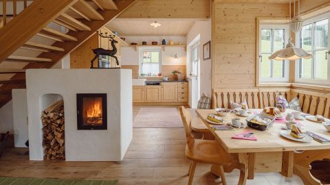 Gemütlicher hölzerner Essbereich mit Frühstücksset, Kamin und gestapeltem Brennholz; die Treppe führt nach oben, die Küche ist im Hintergrund zu sehen.