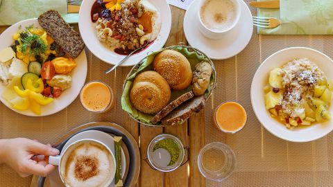 Frühstückstisch mit Kaffee, Saft, Brötchen, Obstsalat, Müsli und Käse auf einem Holztisch, von oben gesehen.