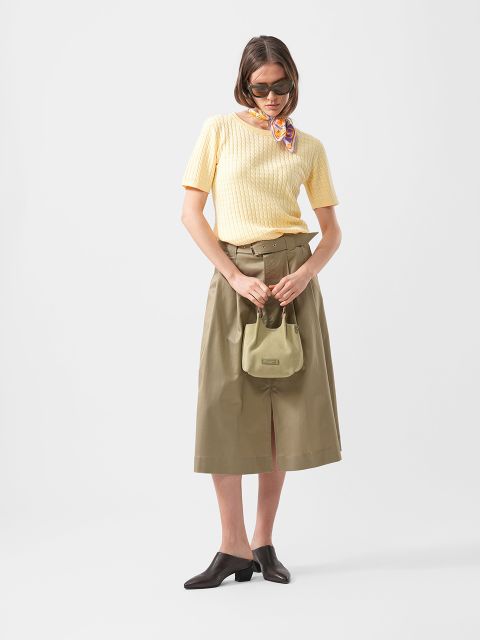 Eine Frau mit Sonnenbrille trägt ein gelbes kurzärmeliges Strickoberteil, einen khakifarbenen Midirock, einen Schal, braune Schuhe und eine kleine grüne Handtasche.