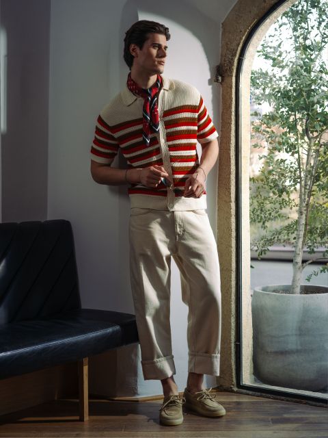 Ein Mann in einem gestreiften Strickpolo, einer cremefarbenen Hose und einer Sonnenbrille posiert am Fenster mit einer Pflanze.