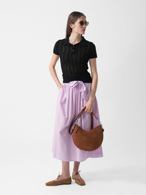 Frau mit Sonnenbrille, die ein schwarzes kurzärmeliges Oberteil, einen hellvioletten Rock und braune Schuhe trägt und eine braune halbmondförmige Handtasche vor einem weißen Hintergrund hält.