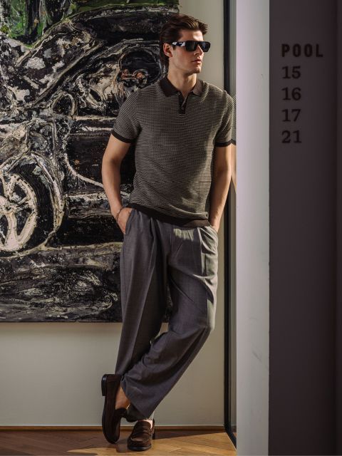 Ein Mann mit Sonnenbrille und gestreiftem Poloshirt lehnt an einer Wand, die Hände in den Taschen.