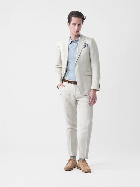 Mann in cremefarbenem Anzug, blauem Hemd, braunem Gürtel und braunen Schuhen, der vor einem schlichten weißen Hintergrund steht und in die Kamera schaut.