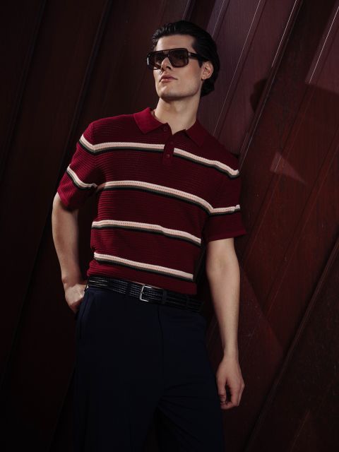 Ein Mann mit Sonnenbrille und einem rot gestreiften Polohemd posiert selbstbewusst mit einer Hand in der Tasche vor einem dunklen, holzgetäfelten Hintergrund.