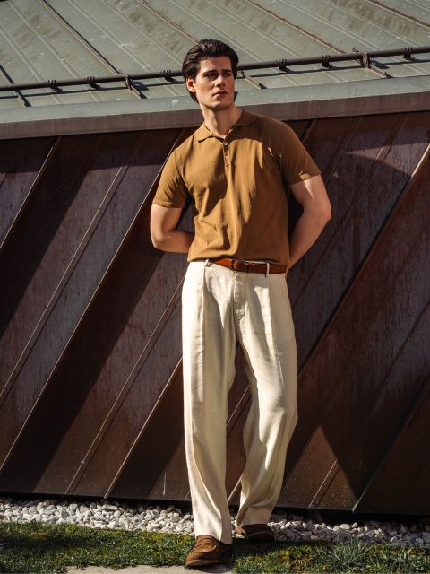 Ein Mann steht im Freien, trägt ein braunes Poloshirt, eine cremefarbene Bundfaltenhose und braune Schuhe und lehnt an einem Metallgebäude im Sonnenlicht.